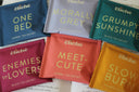 A collection of tea sachets with colorful packaging, each labeled with a different name such as 'Morally Grey', 'Grumpy Sunshine', and 'Meet Cute'.
