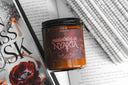 Midwinter in Nyaxia book candle from The Crowns of Nyaxia series surrounded by open books and red poppy