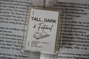 Wax melt packaging for the scent 'tall, dark and fictional' displayed against an open book background