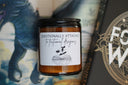 A candle with a label that reads 'Emotionally Attached To Dragons' placed next to a book with a dragon on the cover.