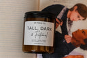 A brown soy candle with a label that reads 'Tall, Dark & Fictional' placed on an open book.